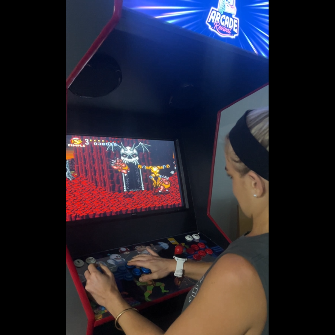 Woman playing Battletoads on a custom-built Arcade Revival machine with illuminated marquee and dual joystick control panel.