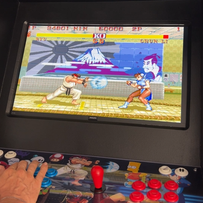 Close-up of an arcade machine running Street Fighter II, with a player controlling Ryu using the blue joystick and buttons on a custom artwork control panel featuring classic characters like Ryu and Bart Simpson.
